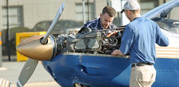 The nose of a plane with the engine exposed and two people working on it