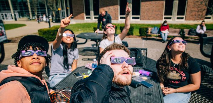 Five students sitting at a table gaze and point at the sun while wearing glasses. 