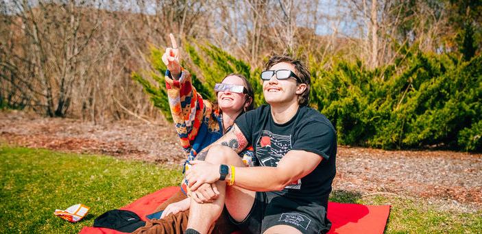 Two students sitting on a blanket gaze and point at the sun while wearing glasses.