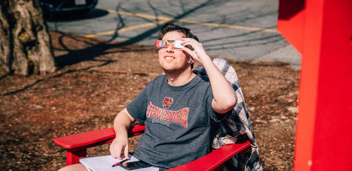 A student sitting in a chair gazes at the sun while wearing eclipse glasses.