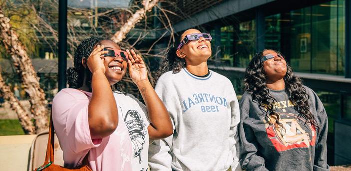 Three students look at the sun while wearing eclipse glasses.